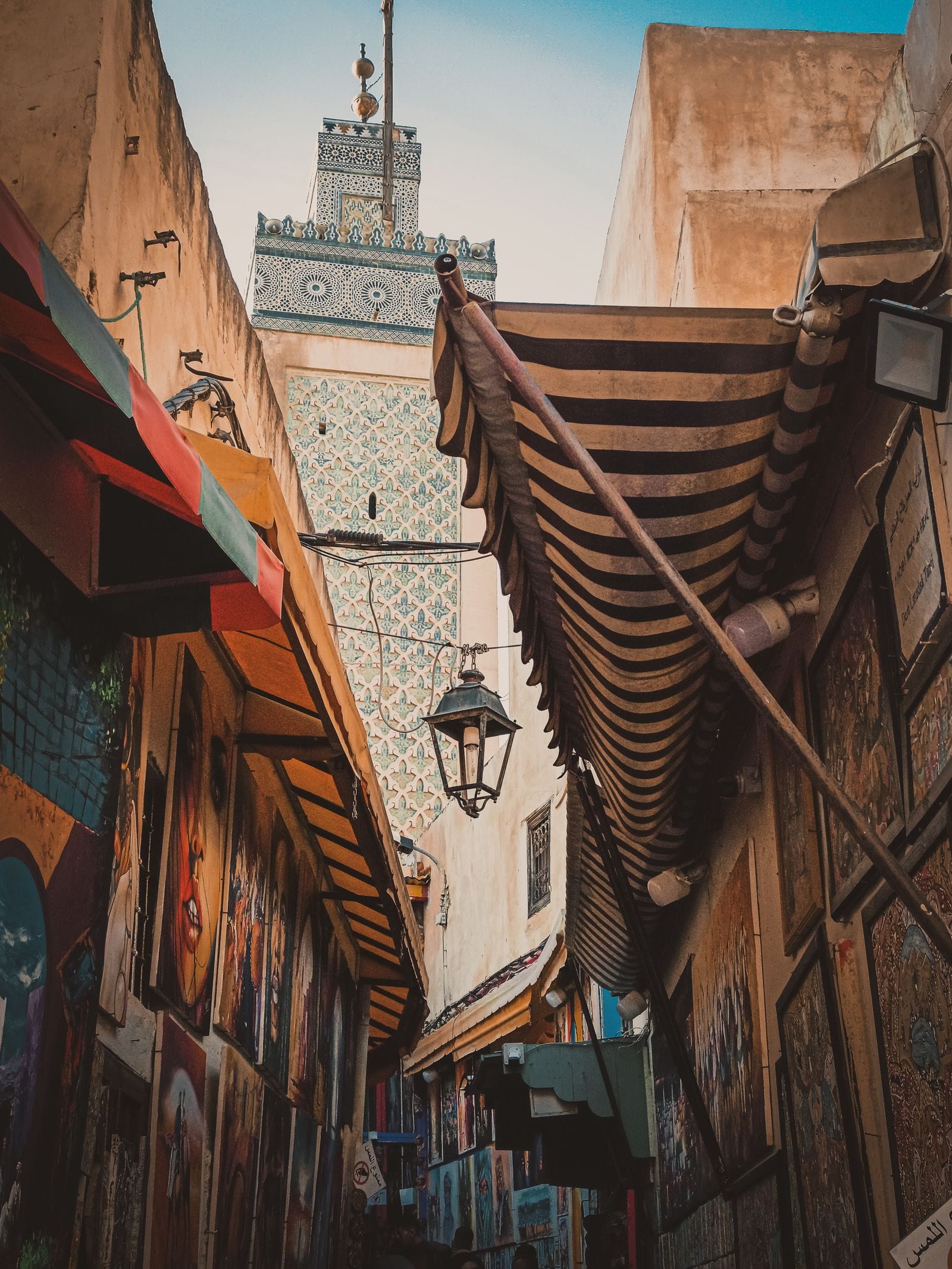A narrow lane in the Fes medina, awnings overhead and the tiled minaret of the Bou Inania Madrasa rising behind the rooftops