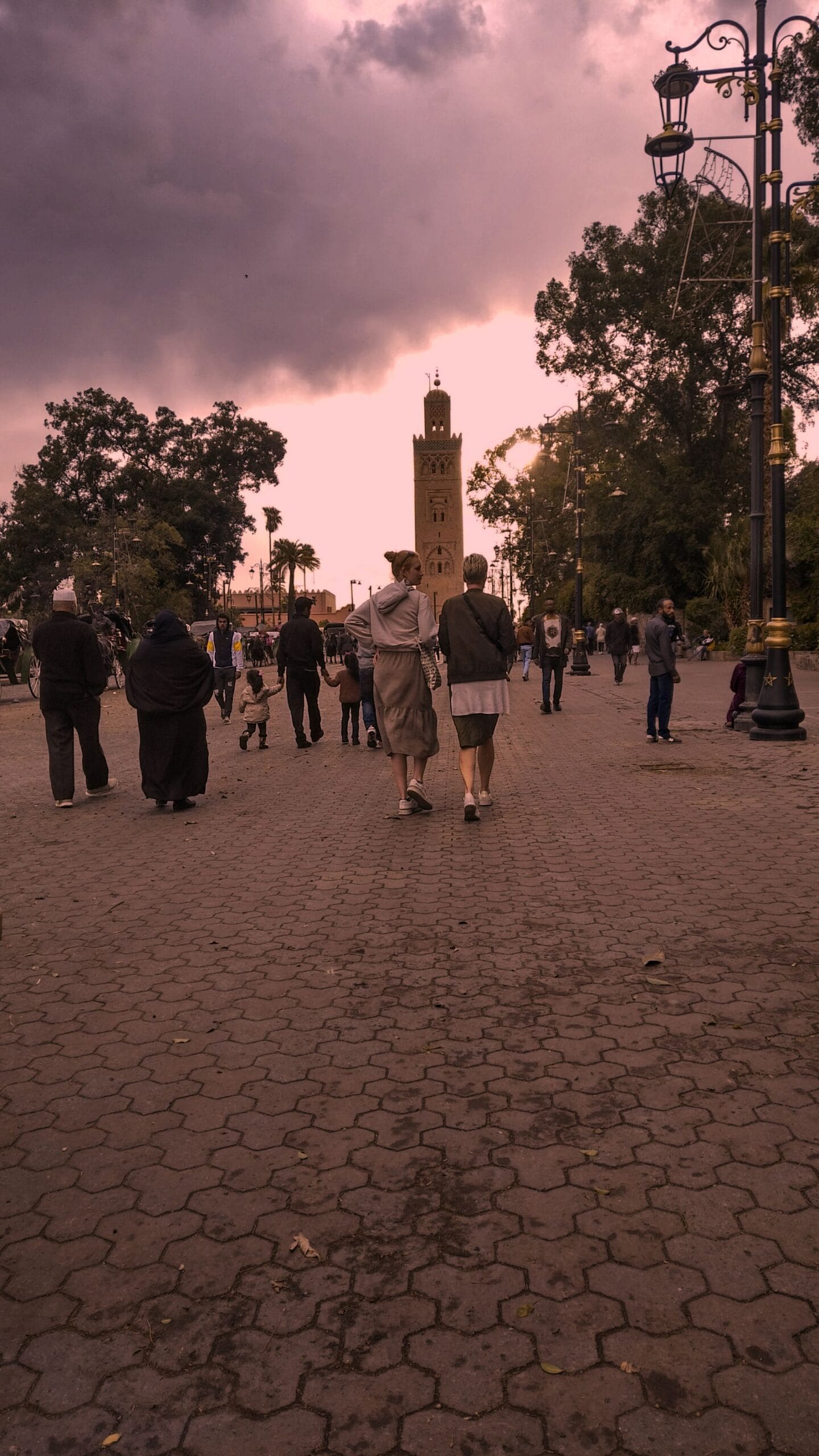 A wide paved esplanade in Marrakech at dusk, people walking, the Koutoubia minaret behind them and the street lamps lit under a heavy pink sky