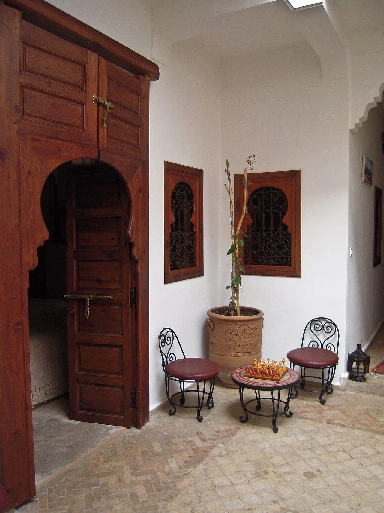 The courtyard corner of a small Marrakech riad guesthouse: carved cedar doorway, iron-grille windows, two chairs and a chess set on a mosaic table