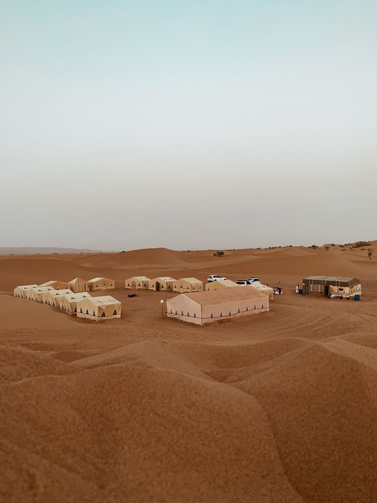 An aerial view of a Sahara camp, about fifteen tents and two 4x4s arranged on low rolling dunes rather than a high dune sea