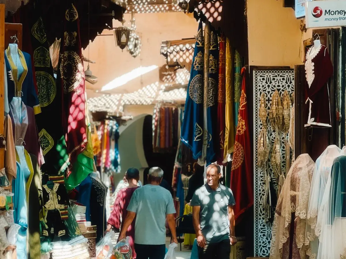 A covered souk alley in Fes lined with embroidered kaftans and a money-transfer sign, the kind of street where nothing carries a price label