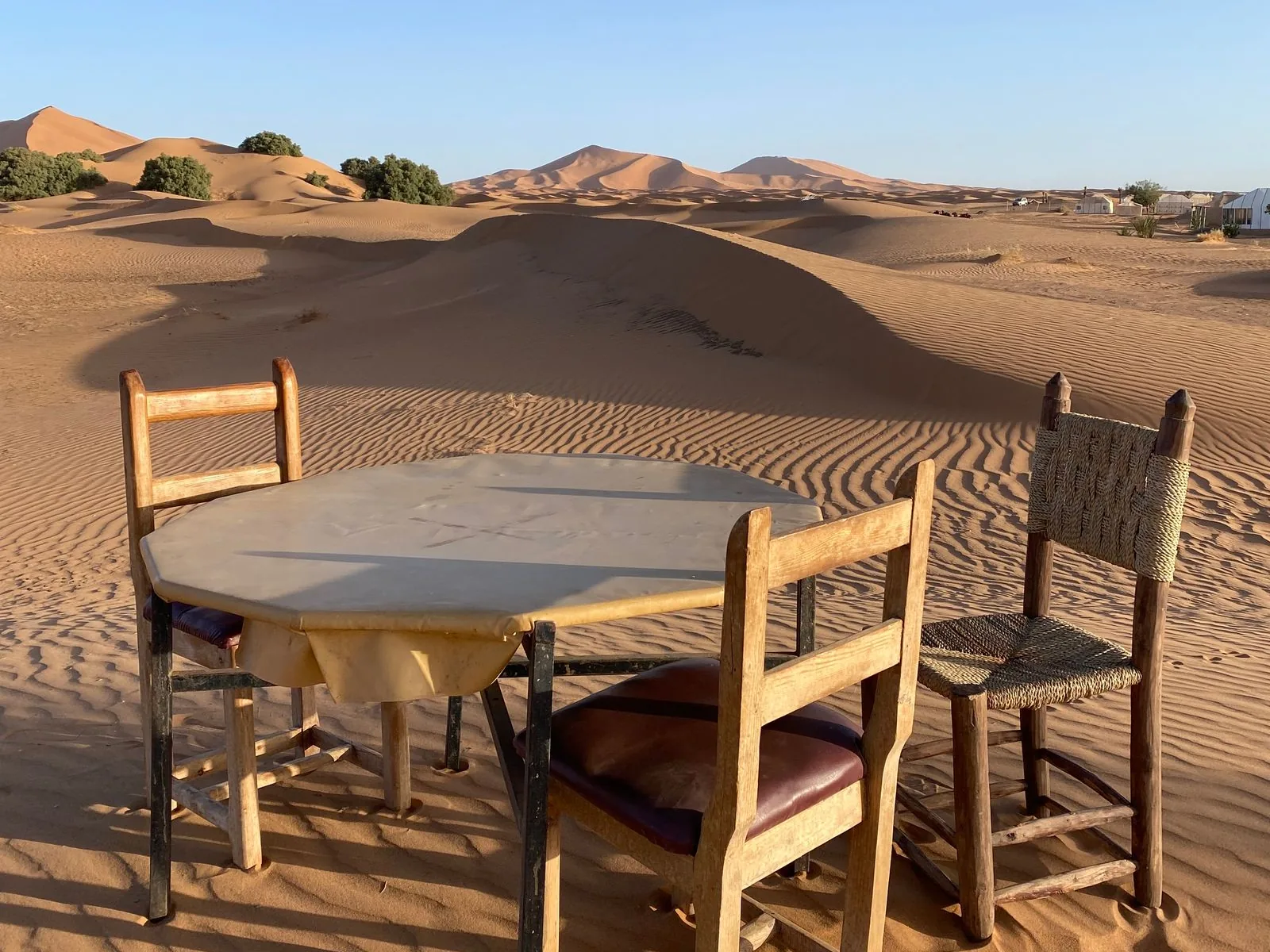 A round table and three wooden chairs set out on the sand at an Erg Chebbi camp, with the dunes and a row of tents behind