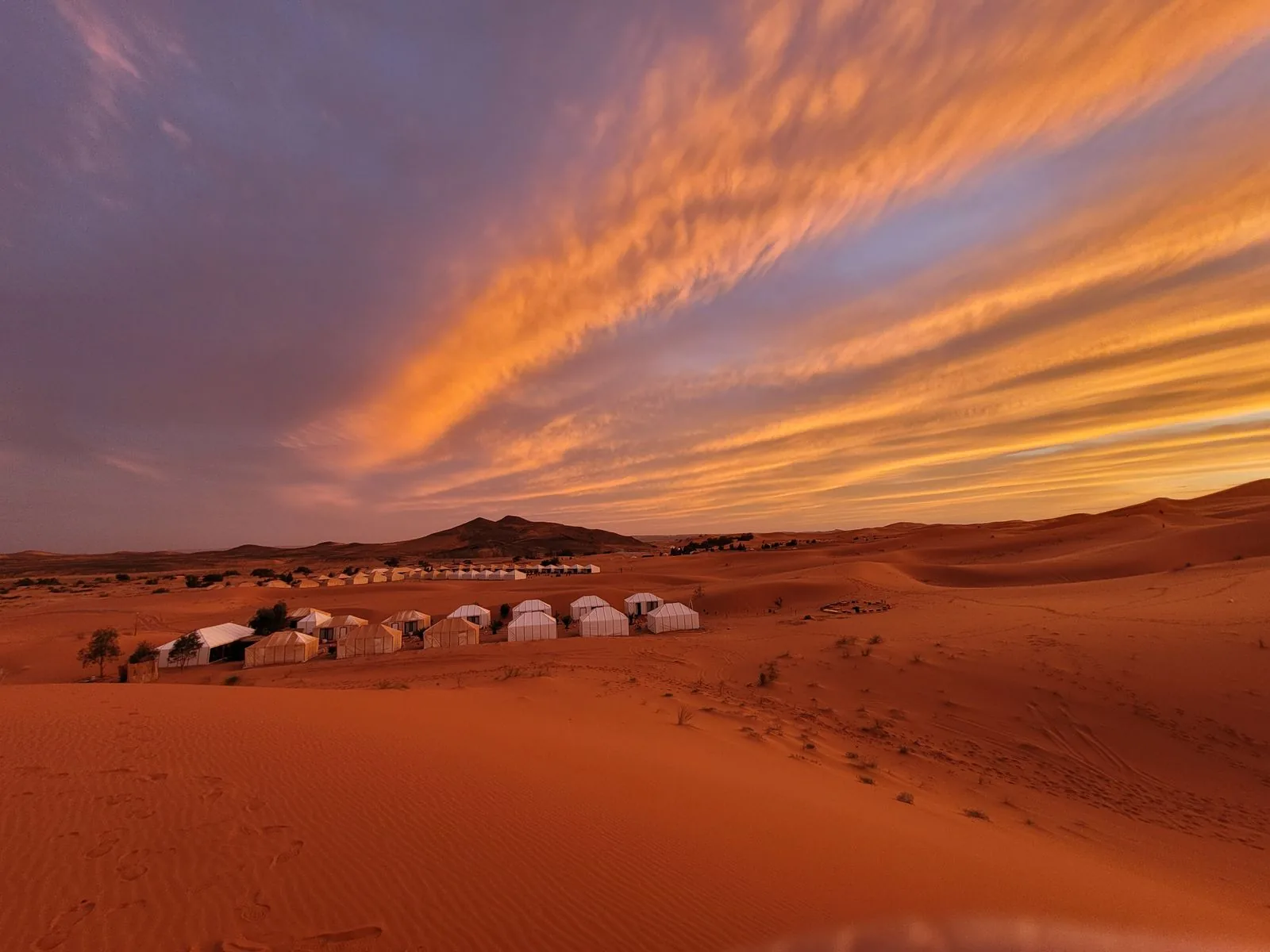 A row of white desert camp tents at Erg Chebbi under a streaked orange dusk sky, the season’s evening light