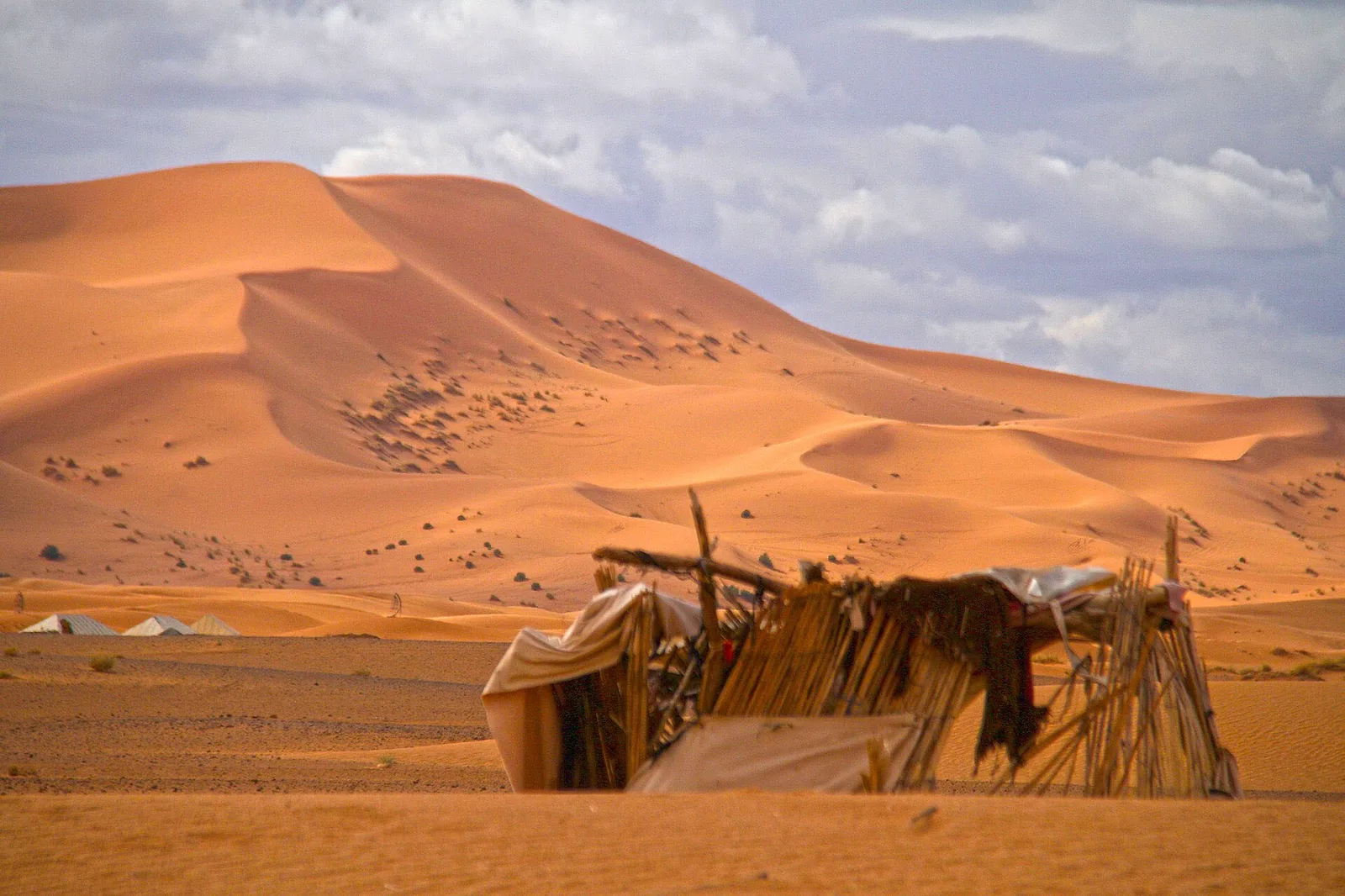 A nomad-style tent of poles and cloth in the foreground with a row of white luxury tents and the Erg Chebbi dunes behind